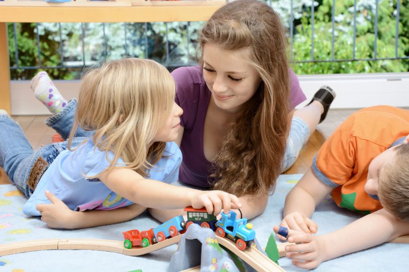 Teenager with two children playing with a wooden train set.