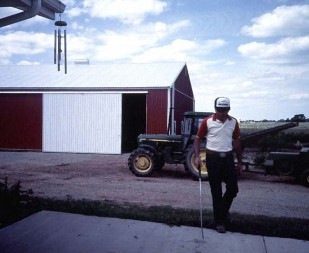 Windchimes hanging from the corner of a strucure and a person walking with a white cane below.