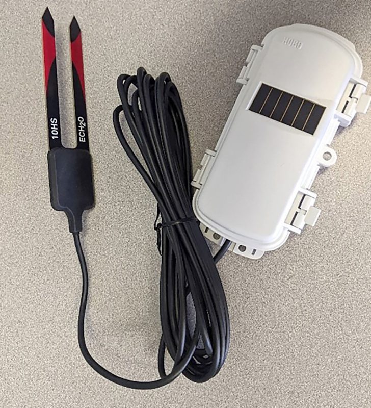 A photograph of a volumetric water content sensor with two red and black prongs attached via a black cord to a white remote data collection device with a solar panel.