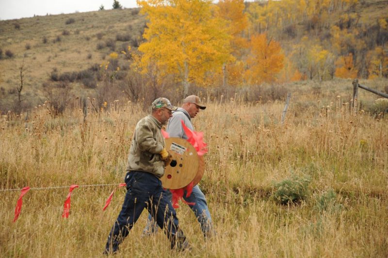 Two men walking in a field unrolling a large wooden spool of wire, carrying it about waist high. Bright red-orange elongated rectangular flags hang from the wire about every 18 inches.