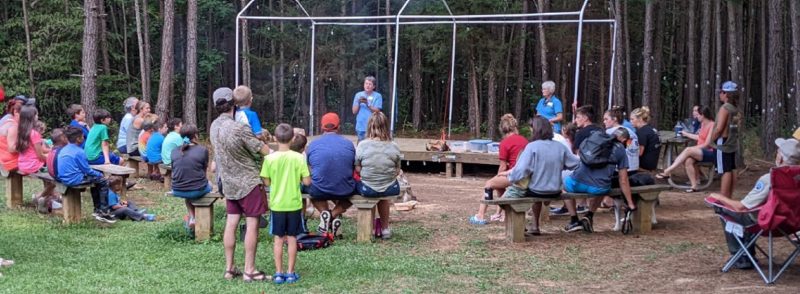 Two VMN volunteers stand under the frame of a shelter, one holding a turtle, and face a group of onlookers of all ages seated on benches