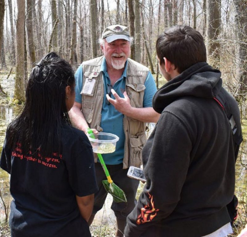 A man holding a water skimmer and a plastic container, talks with two young people who have their backs to the camera