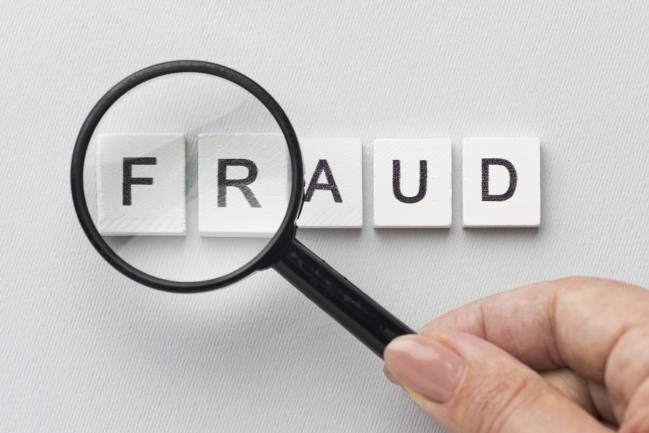 A hand holding a magnifying glass over the word 'FRAUD' spelled out on tiles. 