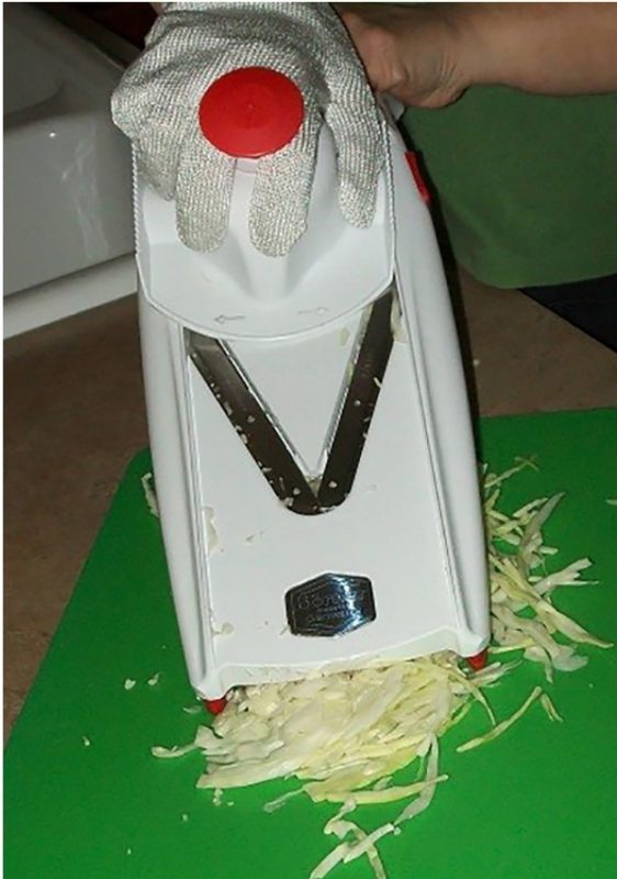 A person wearing a cut-resistant glove uses a Mandolin on a cutting board to shred cabbage.	