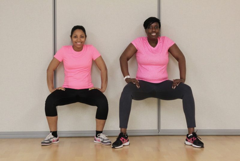Two women sitting against the wall with their toes slightly turned away from them and bended their knees to lower into a squate.