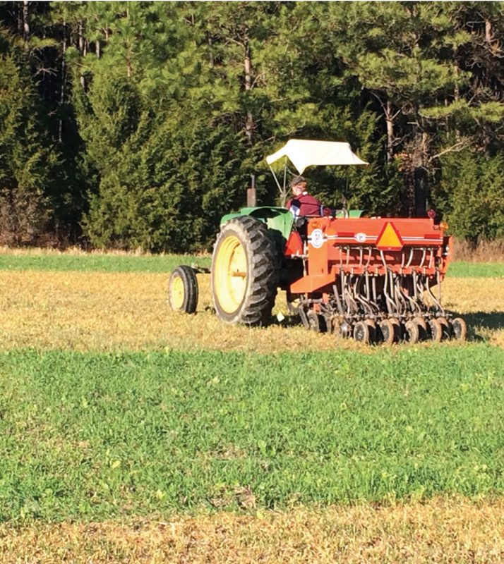 A person driving a tractor across a partially vegetated field, with a forested background.
